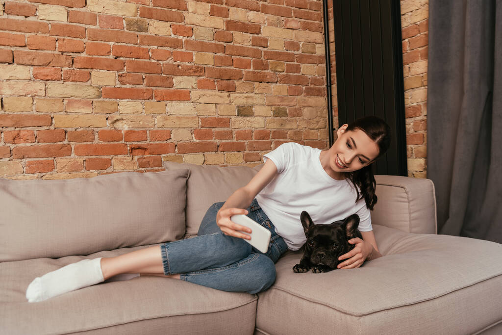 happy young woman taking selfie with cute french bulldog in living room  - Photo, Image