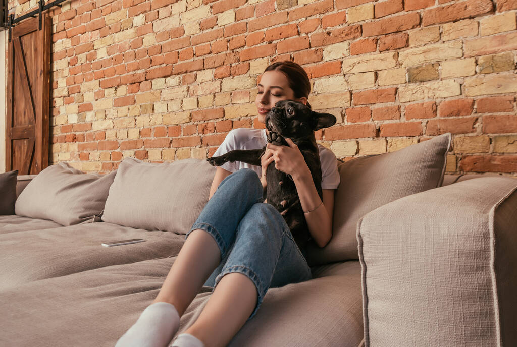 glückliches Mädchen hält süße französische Bulldogge in den Armen, während sie auf dem Sofa in der Nähe von Smartphone sitzt  - Foto, Bild