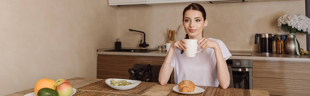 horizontales Bild einer attraktiven Frau, die eine Tasse Kaffee in der Nähe von leckerem Frühstück und französischer Bulldogge hält  - Foto, Bild