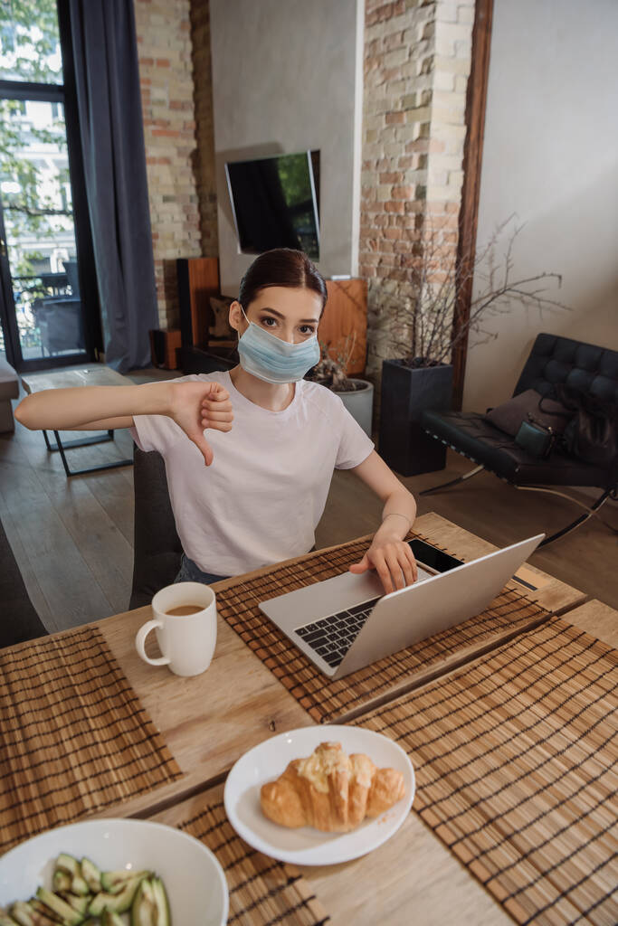 młody freelancer w masce medycznej pokazując kciuk w dół w pobliżu laptopa - Zdjęcie, obraz