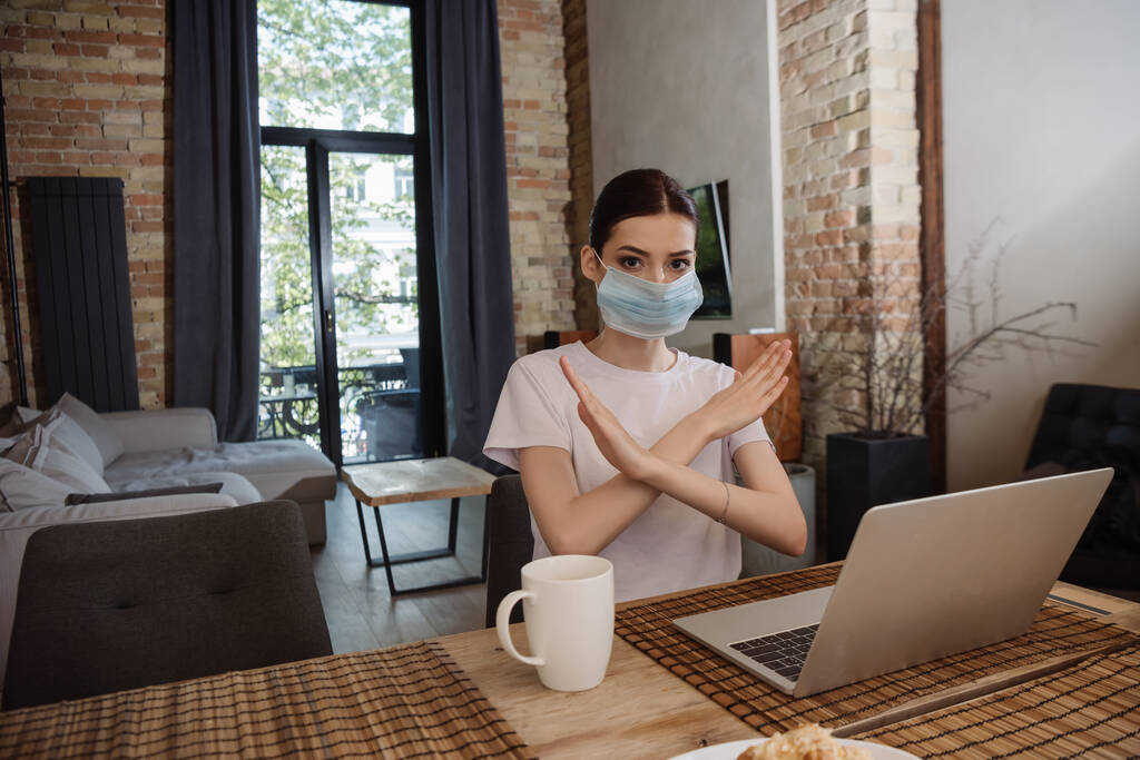 joven freelancer en máscara médica mostrando stop sign cerca de laptop
 - Foto, Imagen