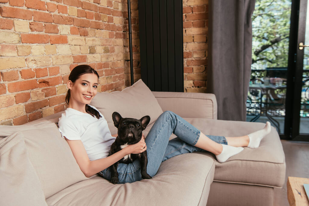 fröhliche junge Frau sitzt auf Sofa mit schwarzer französischer Bulldogge  - Foto, Bild