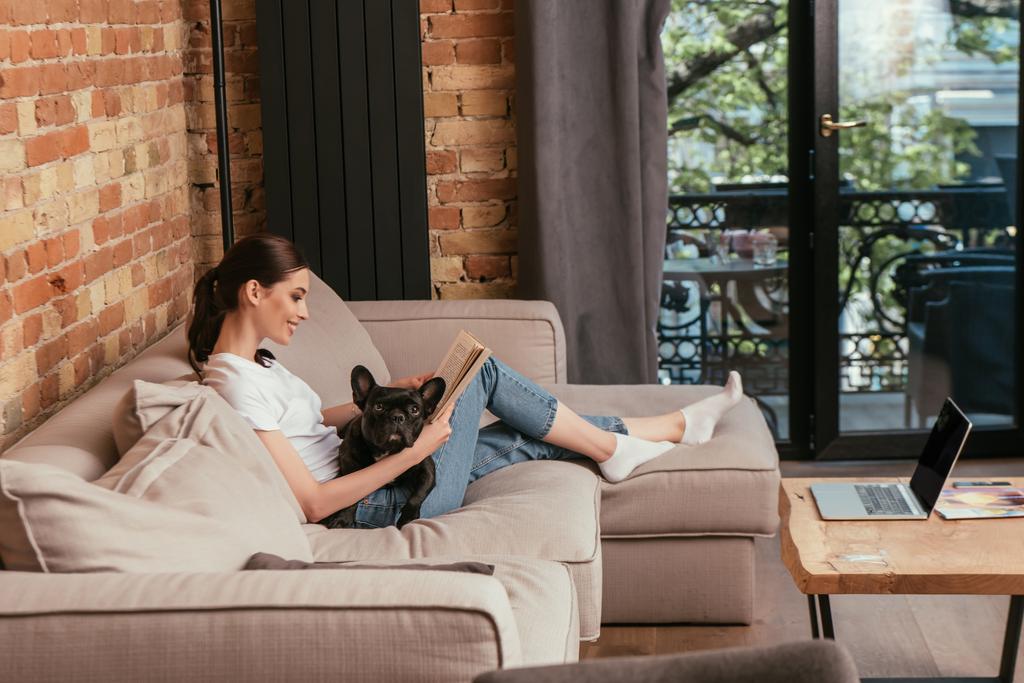 вид сбоку на счастливую молодую женщину, читающую книгу рядом с черным французским бульдогом и ноутбуком с бланковым экраном
  - Фото, изображение
