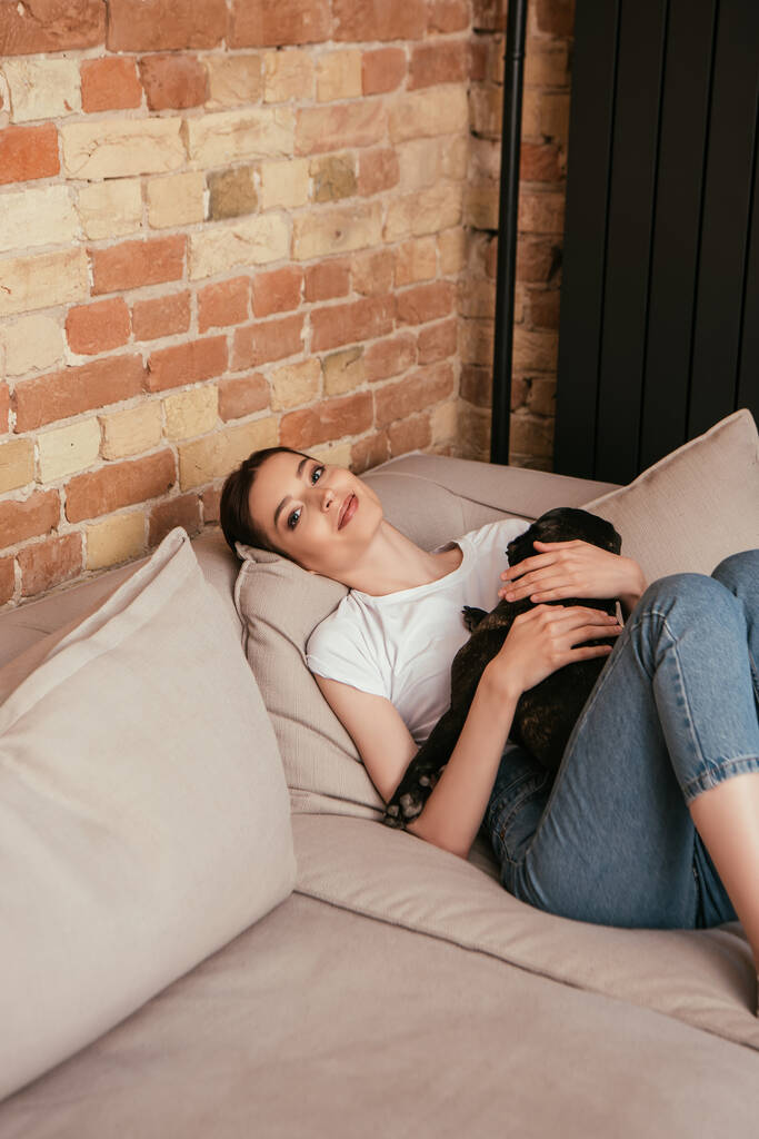 happy woman hugging french bulldog and looking at camera in living room - Photo, Image