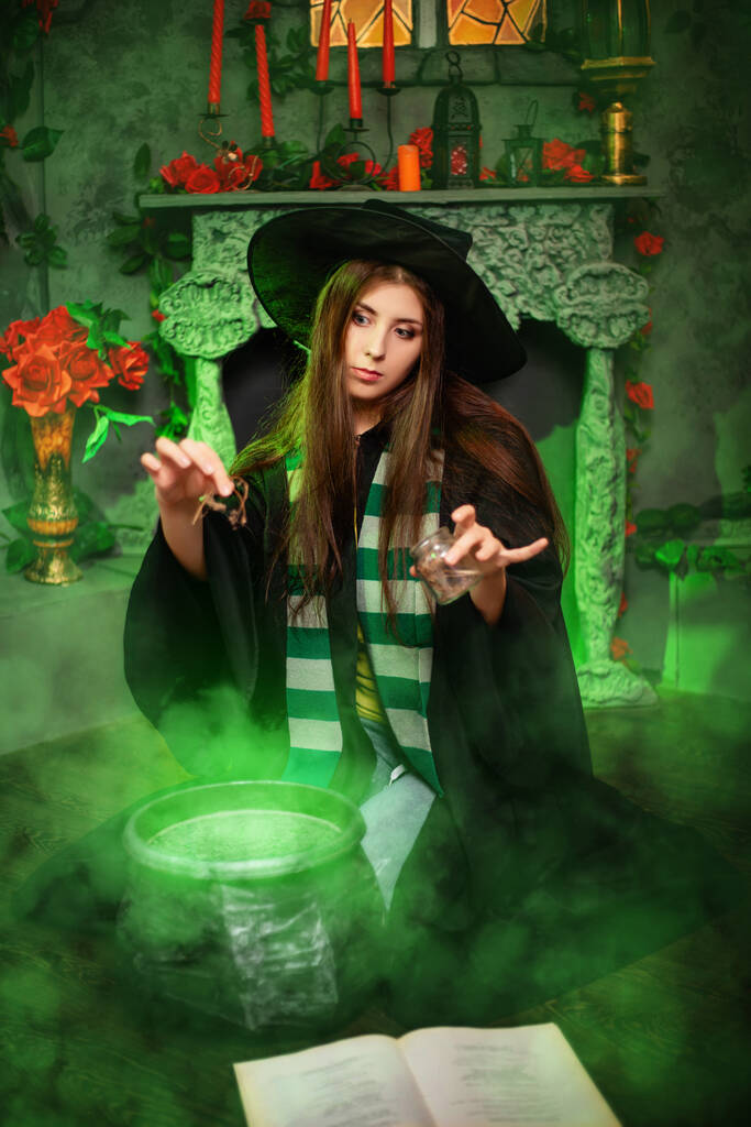 Vonzó lány egy fekete kalapba és köpenybe öltözött boszorkány képében hozzávalót dob egy üstbe egy közelgő bájitallal, egy varázslatos, zöld füstben ülve. Halloween boszorkány koncepció. - Fotó, kép