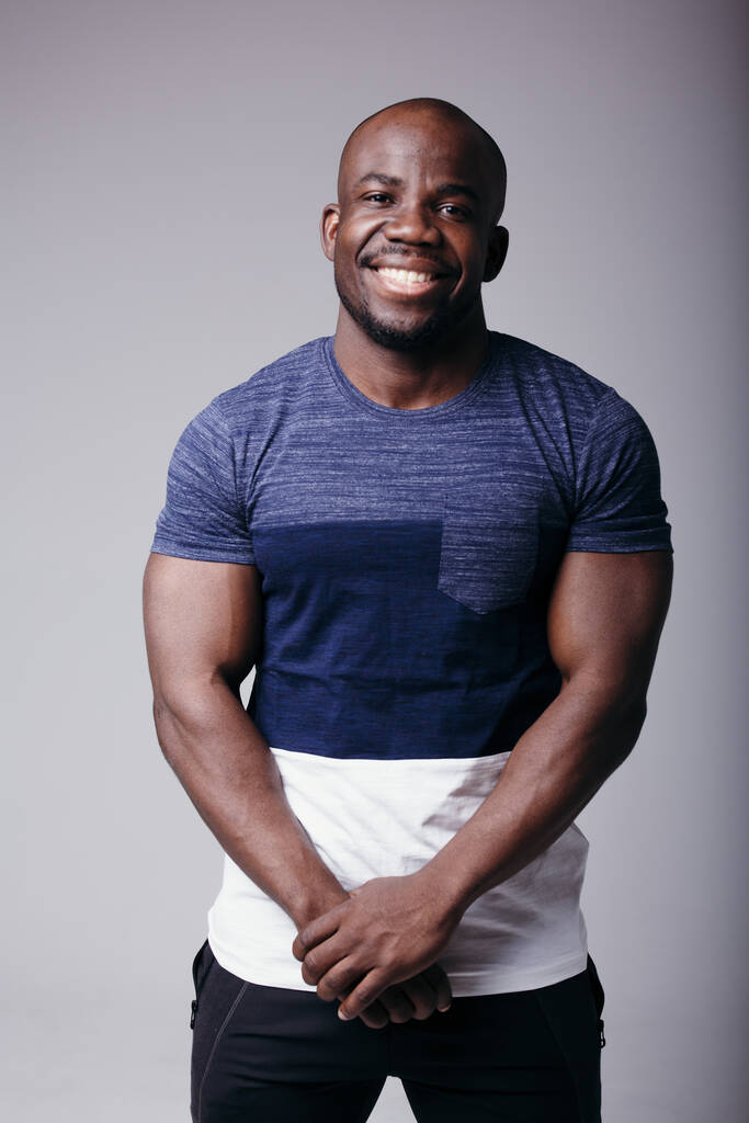 Retrato de un hombre negro con una camiseta de color sosteniendo sus manos cerca de su estómago y sonriendo a la cámara en el estudio
 - Foto, Imagen