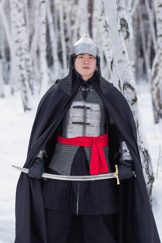   チェーンメールで中世の戦士,ヘルメット,冬の白樺の森の背景に彼の手にサーベルと黒のマント. - 写真・画像
