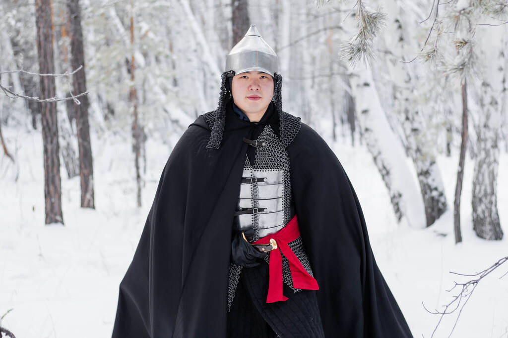 Mittelalterlicher Krieger in Rüstung, Helm, schwarzem Mantel mit Säbel in den Händen, der im Winter im Wald spaziert. - Foto, Bild