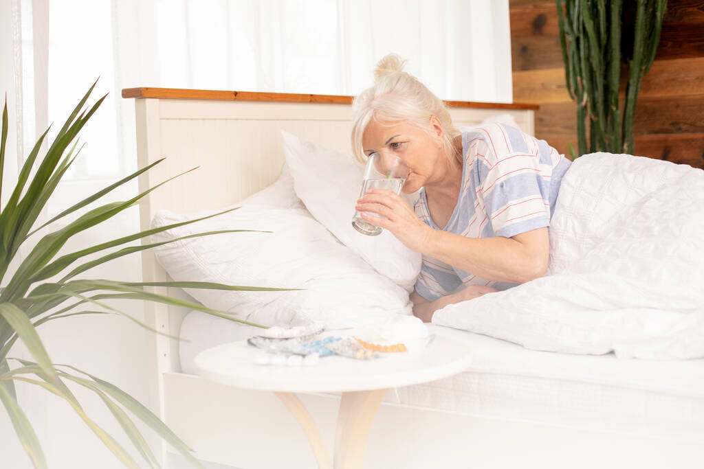 Εποχιακή γρίπη. Άρρωστη ηλικιωμένη γυναίκα με πυρετό και μύτη που τρέχει στο κρεβάτι, στο σπίτι. - Φωτογραφία, εικόνα