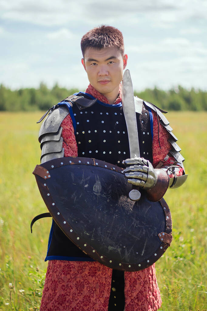 Steppe warrior in Mongolian armor of the 14th century in the field against the background of the forest and the blue sky. With a shield and a sword in his hands. Asian soldier nomad. - Photo, Image