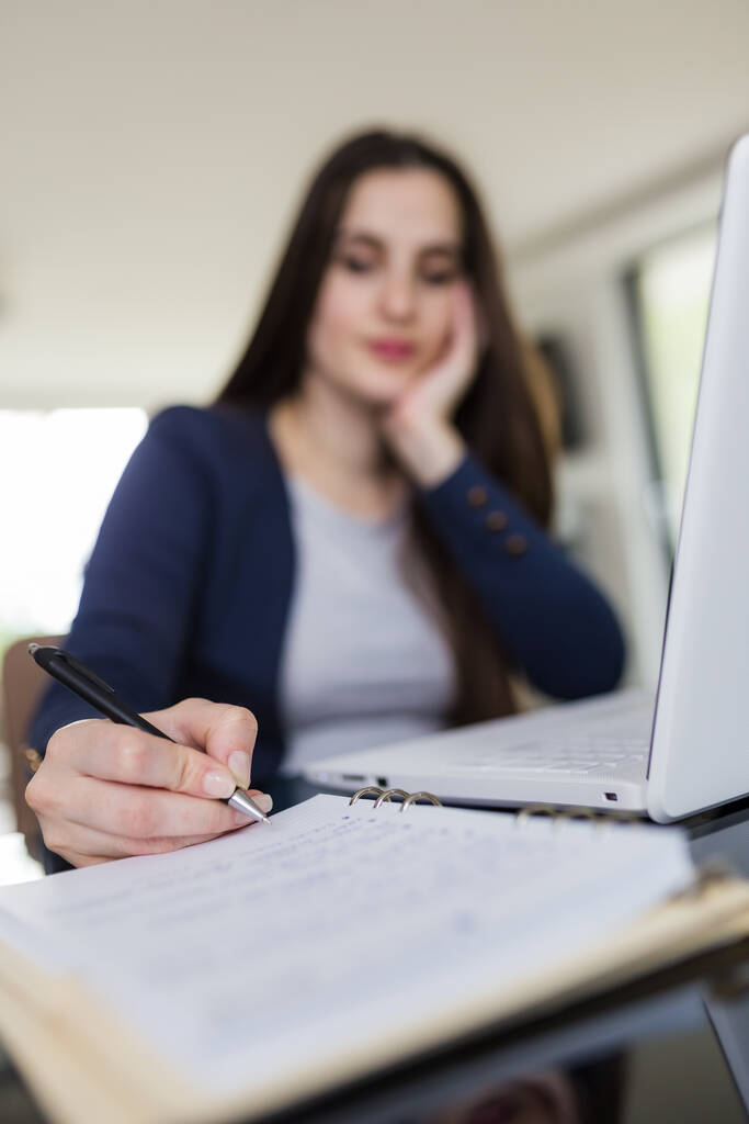 Masasında dizüstü bilgisayarının önünde oturan ve not alan genç bir kadının portresi. E-öğrenme kavramı. - Fotoğraf, Görsel