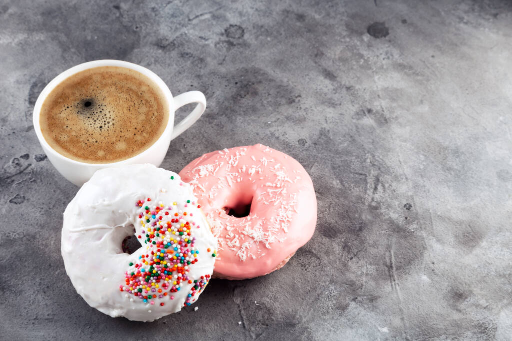 Valkoinen kuppi kahvia ja kaksi donitsia valkoinen ja vaaleanpunainen kuorrutus ja monivärinen sokeri leivonnaiset päällä harmaalla pohjalla. Kopioi tila - Valokuva, kuva
