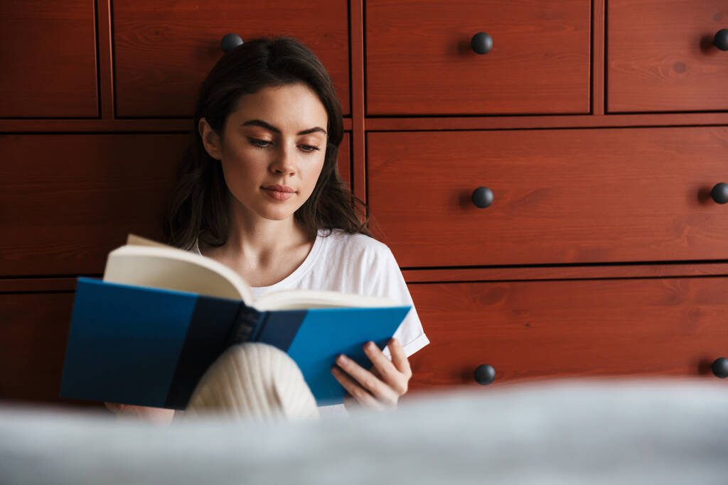 Atractiva joven morena leyendo un libro mientras está sentada en el suelo y apoyada en un armario
 - Foto, Imagen
