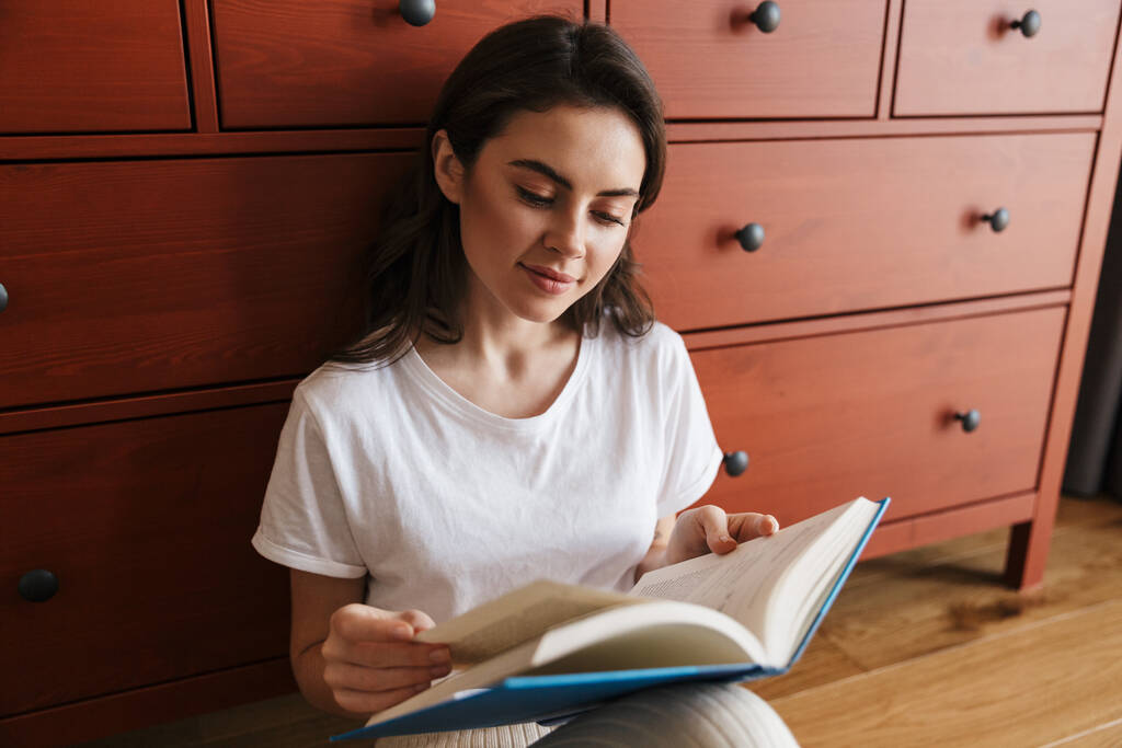 Aantrekkelijke glimlachende jonge brunette vrouw het lezen van een boek terwijl zitten op een vloer en leunen op een kast - Foto, afbeelding