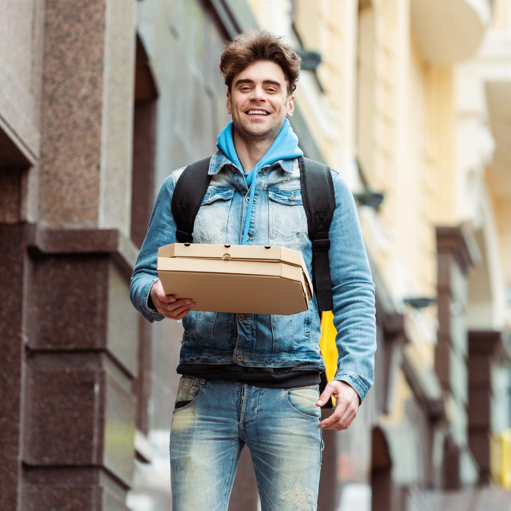 Όμορφος κούριερ χαμογελάει στην κάμερα ενώ κρατάει κουτιά πίτσας στον αστικό δρόμο  - Φωτογραφία, εικόνα