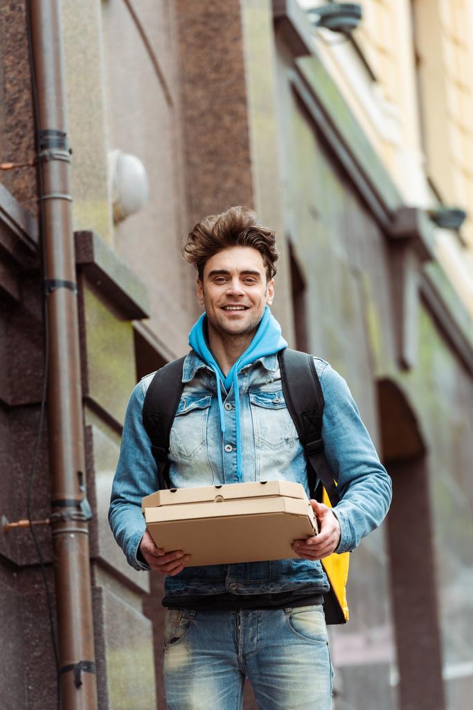 Przystojny kurier uśmiechający się do kamery trzymając pudełka po pizzy na miejskiej ulicy  - Zdjęcie, obraz