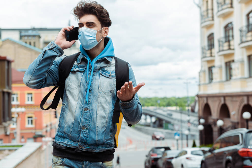 Auslieferer in medizinischer Maske spricht auf Smartphone nahe Straße in der Stadt  - Foto, Bild