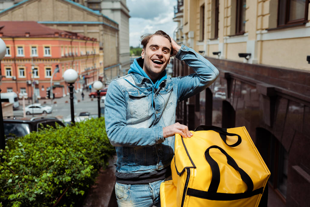 Positiver Kurier lächelt in die Kamera, während er Thermobeutel in der Stadtstraße hält  - Foto, Bild
