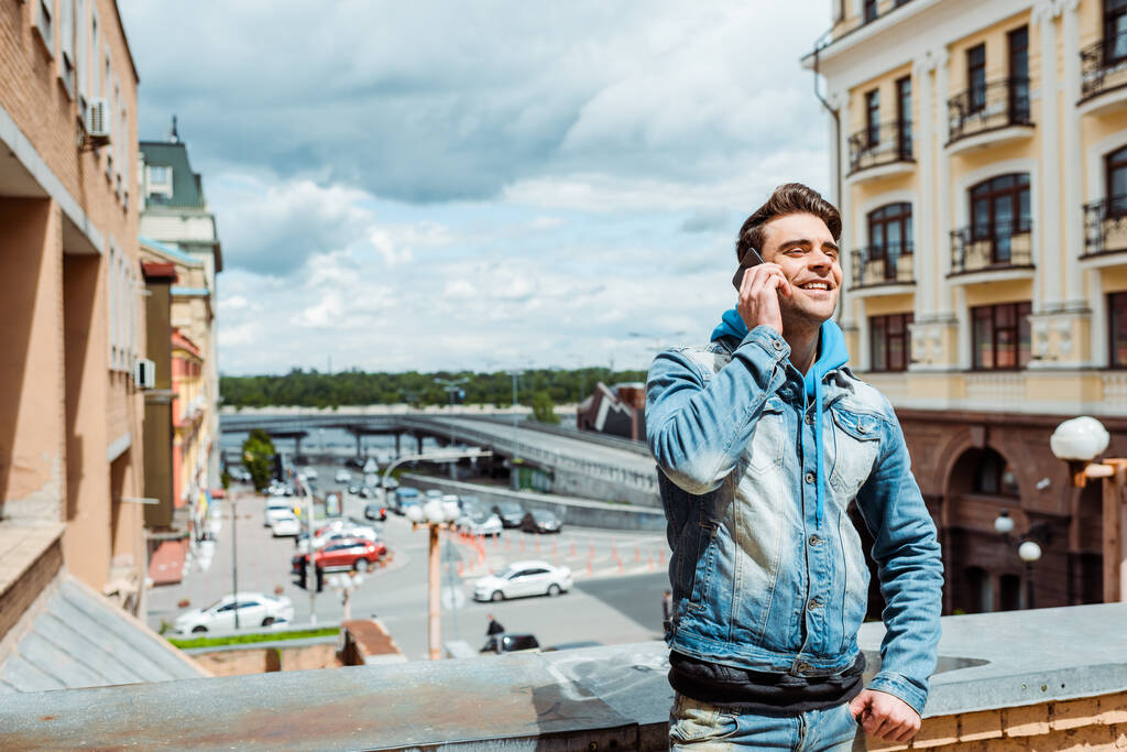 Schöner Mann lächelt, während er auf dem Smartphone mit wolkenverhangenem Himmel und Stadtstraße im Hintergrund spricht  - Foto, Bild
