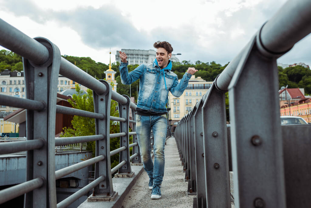 Selective focus of cheerful man in earphones showing yeah gesture while walking on bridge  - Photo, Image