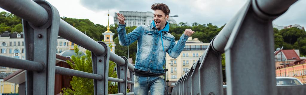 Cultivo panorámico de hombre positivo en auriculares mostrando sí gesto mientras camina sobre puente
  - Foto, Imagen