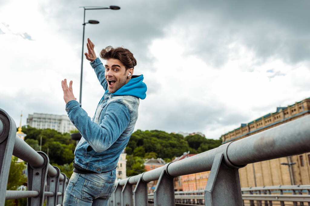 Vista lateral do homem feliz em fones de ouvido olhando para a câmera enquanto caminhava na rua urbana
   - Foto, Imagem