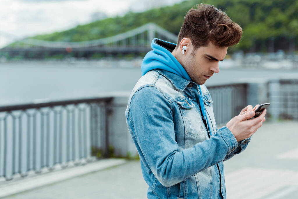 Nuori mies käyttää kuulokkeita ja kännykkää kaupunkien kadulla    - Valokuva, kuva