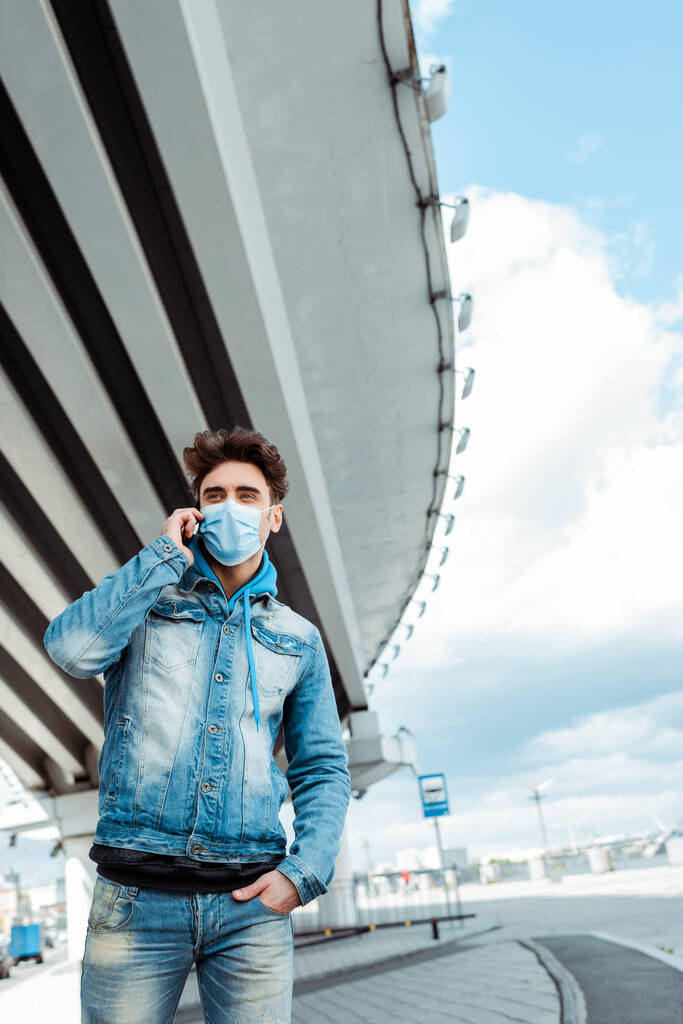 Άντρας με ιατρική μάσκα μιλάει στο κινητό με γέφυρα και συννεφιασμένο ουρανό στο παρασκήνιο  - Φωτογραφία, εικόνα
