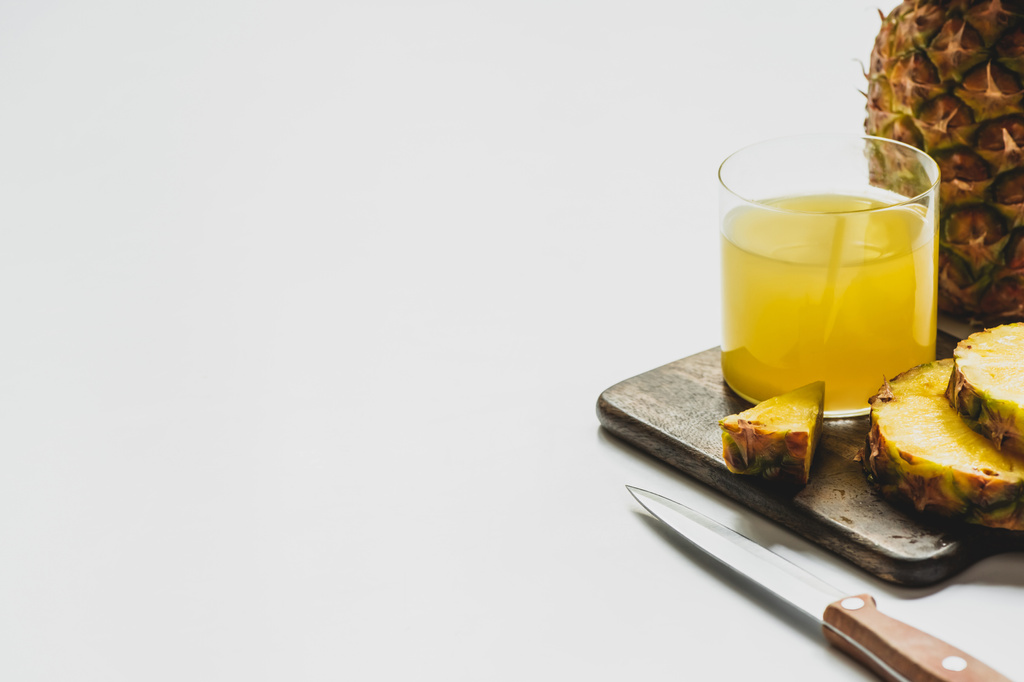 świeży sok ananasowy w szkle w pobliżu pokrojone pyszne owoce na desce do krojenia drewna z nożem na białym tle - Zdjęcie, obraz