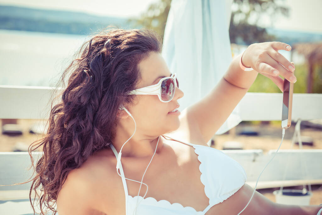 Closeup young beautiful happy, excited smiling woman looking on mobile cell phone taking selfie, listening to music outdoors on sun-bed at sea lake background. Positive facial expression human emotion - Photo, Image