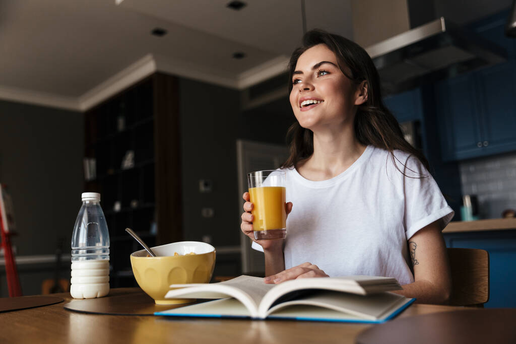 Atraktivní usmívající se mladá brunetka, která čte knihu, zatímco má zdravou snídani, zatímco sedí u kuchyňského stolu a drží sklenici pomerančového džusu - Fotografie, Obrázek