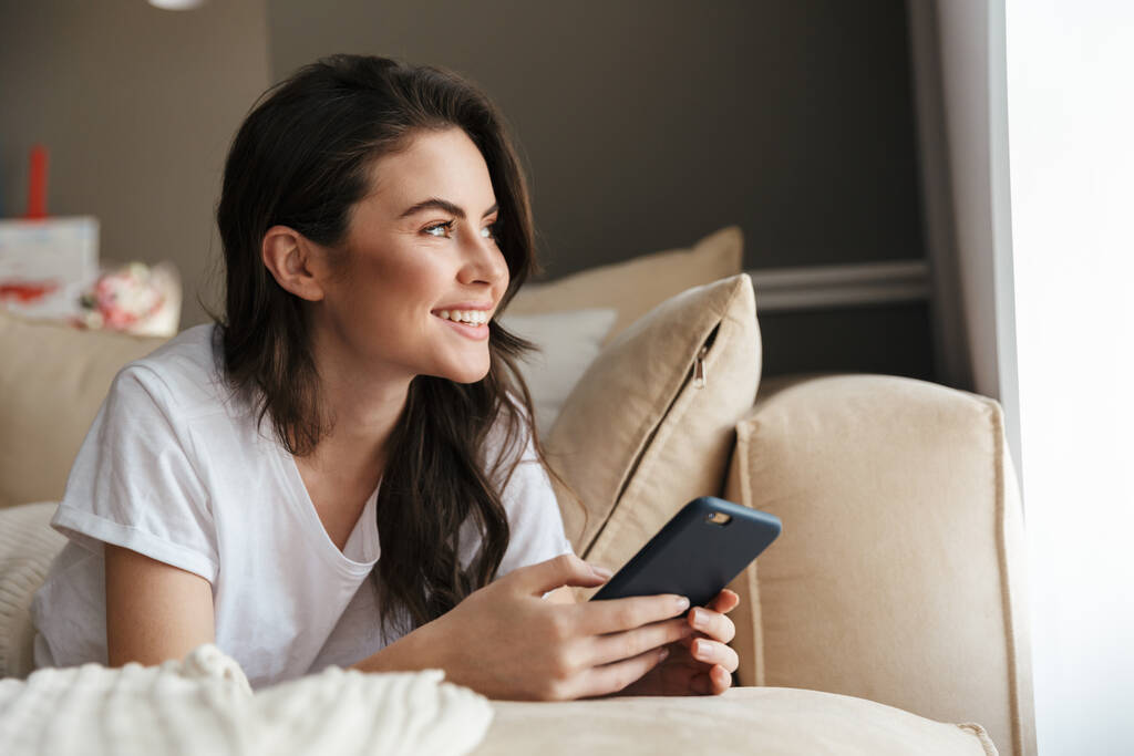 Krásná usmívající se mladá brunetka žena relaxující na gauči doma, pomocí mobilního telefonu - Fotografie, Obrázek