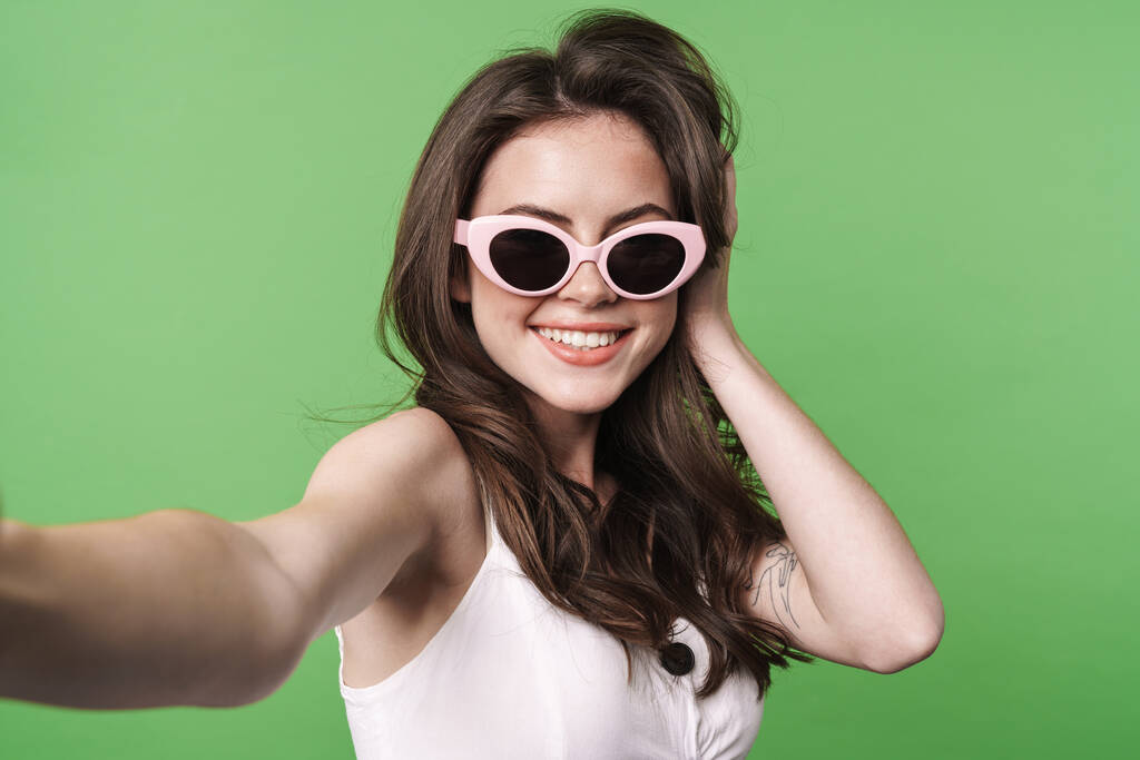 Kép vicces vonzó nő napszemüvegben mosolyog, és hogy szelfi fotó elszigetelt zöld háttér - Fotó, kép