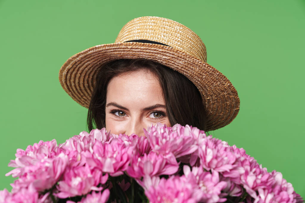 Image closeup of cheerful pretty woman in straw hat holding flowers isolated over green background - Photo, Image