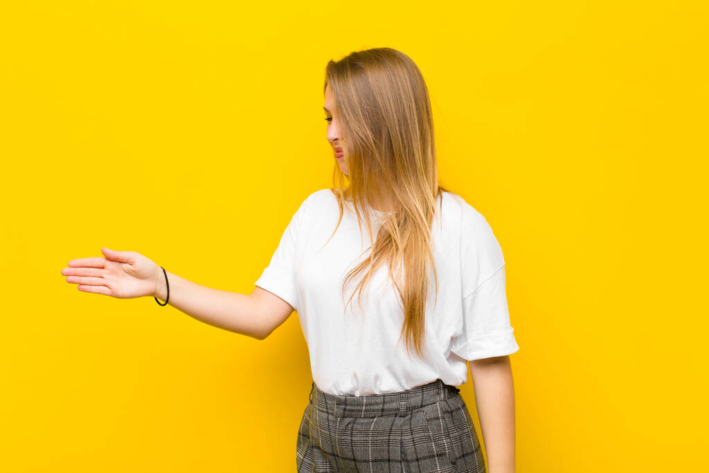 young blonde woman smiling, greeting you and offering a hand shake to close a successful deal, cooperation concept against orange wall - Photo, Image