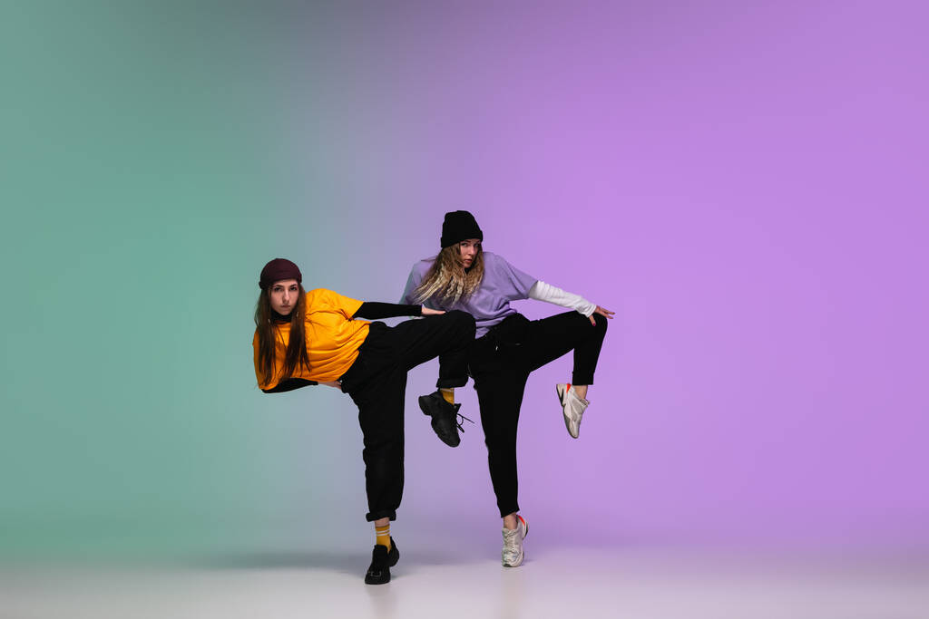 Meisjes dansen hip-hop in stijlvolle kleding op gradiënt achtergrond in danszaal in neon licht. - Foto, afbeelding