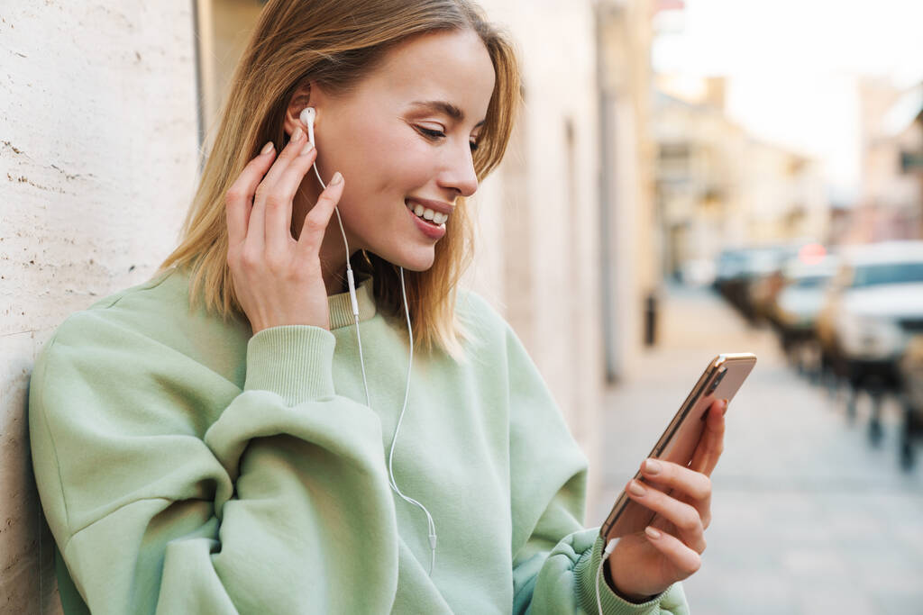 Ritratto di giovane donna sorridente che utilizza auricolari e telefono cellulare mentre cammina per strada
 - Foto, immagini