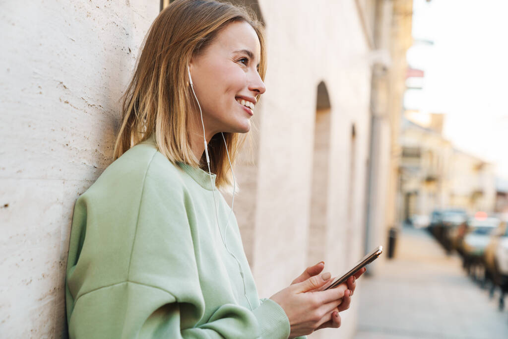 Retrato de una joven sonriente usando auriculares y teléfono móvil mientras camina por la calle de la ciudad
 - Foto, Imagen