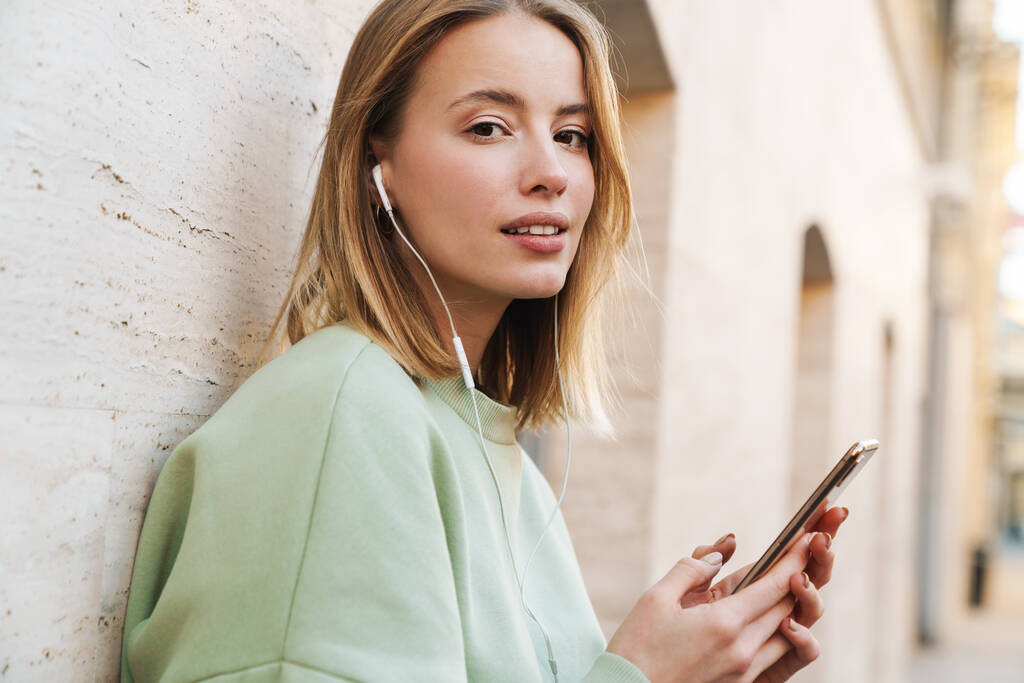 Портрет красивої молодої жінки з використанням навушників та мобільного телефону під час прогулянки по міській вулиці
 - Фото, зображення