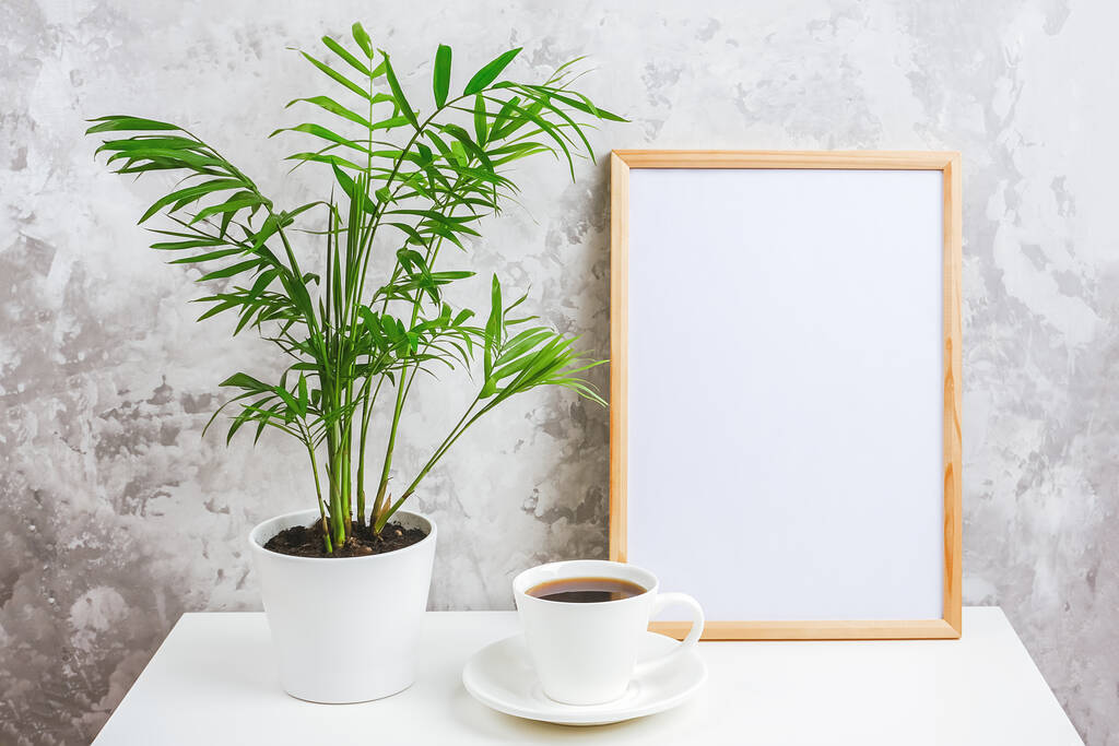 Dřevěný vertikální rám s bílou prázdnou kartou, šálkem kávy a zeleným exotickým květem palmy v květináči na stole na šedém betonovém pozadí. Šablona Mockup pro váš design, text. - Fotografie, Obrázek