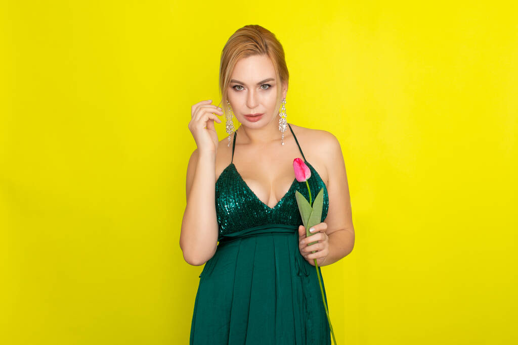 Blondynka w zielonej sukni wieczorowej trzymając jeden tulipan w dłoniach na żółtym tle - Zdjęcie, obraz