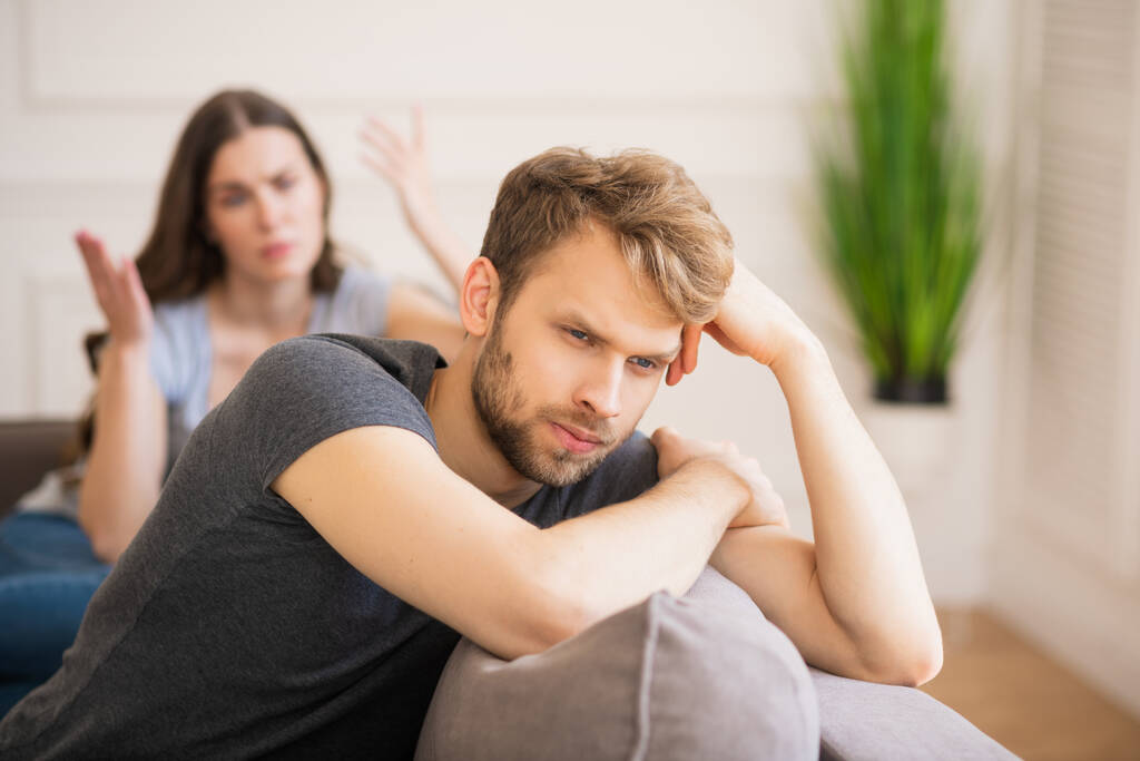 Junger bärtiger Mann sitzt mit deprimiertem Blick da, während seine Frau spricht - Foto, Bild