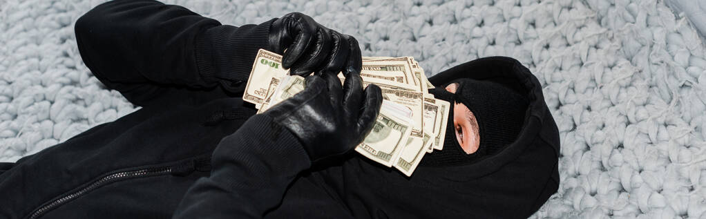 Panoramatický záběr zloděje v balaklavě držící peníze v blízkosti obličeje na posteli  - Fotografie, Obrázek