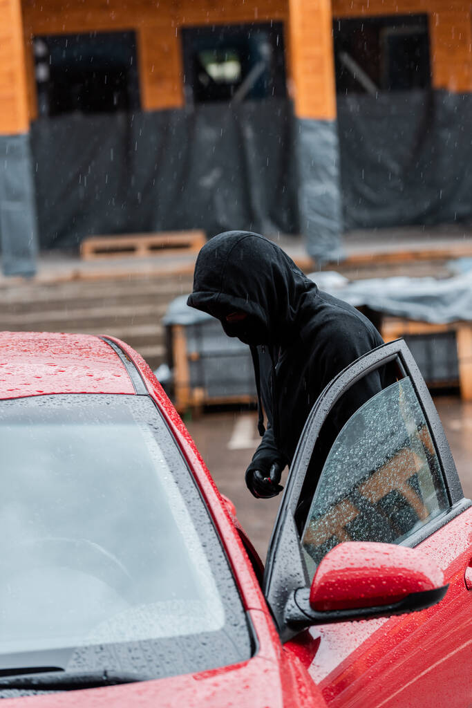 Vista lateral del hombre en pasamontañas y guantes de cuero de pie cerca del coche con la puerta abierta durante el robo
  - Foto, Imagen