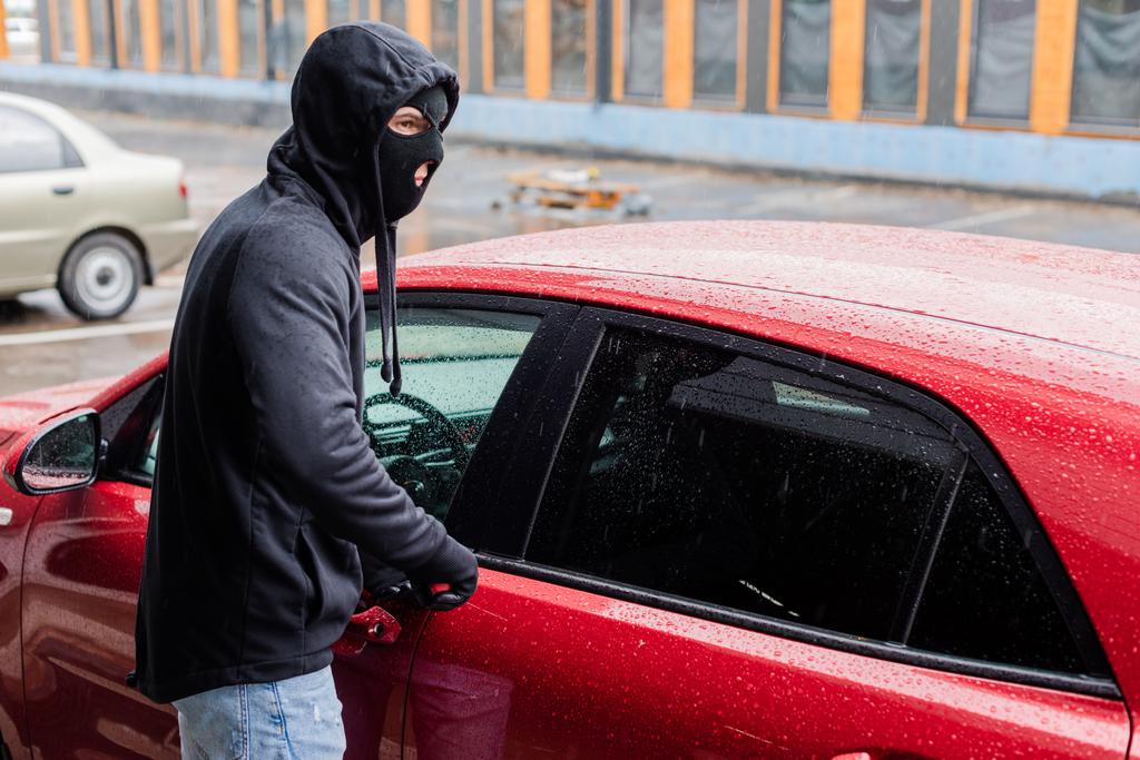 Räuber mit Sturmhaube und Lederhandschuhen schaut weg, während er Auto mit Schraubenzieher auf Stadtstraße öffnet  - Foto, Bild