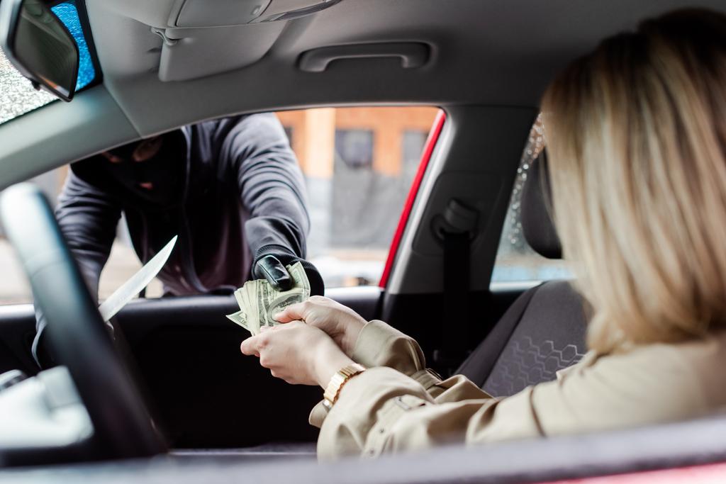 Επιλεκτική εστίαση της γυναίκας δίνοντας μετρητά στο ληστή με μαχαίρι κοντά στο αυτοκίνητο  - Φωτογραφία, εικόνα
