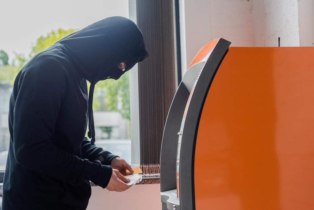 Vue latérale du voleur masqué tenant des cartes de crédit près d'un guichet automatique
 - Photo, image