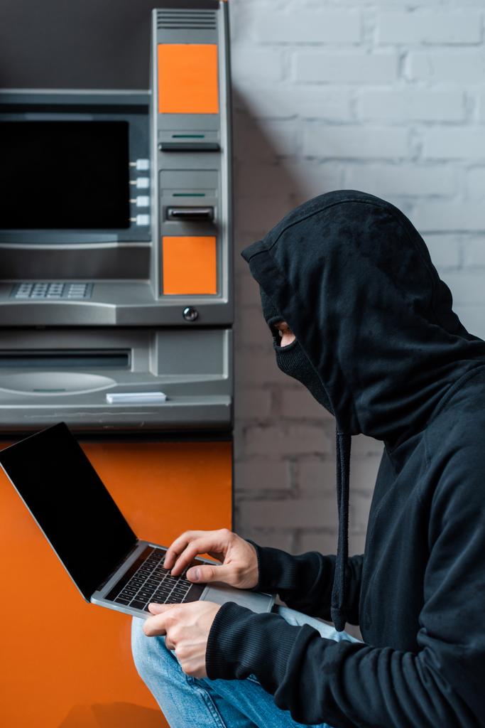 Vista lateral do hacker em balaclava usando laptop perto de ATM
  - Foto, Imagem