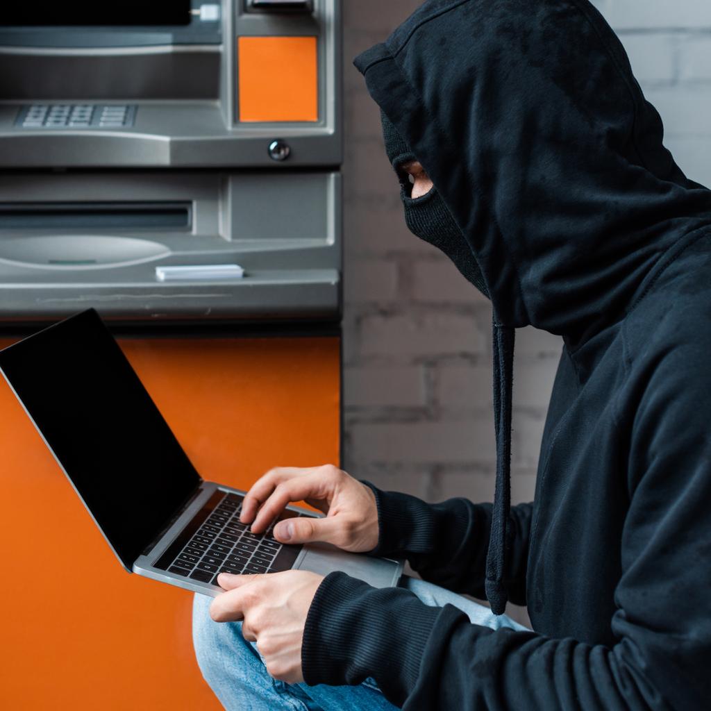 Vista lateral do hacker em balaclava usando laptop perto de caixa automática
 - Foto, Imagem