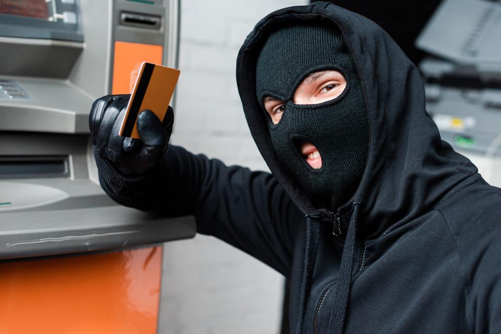 Kar maskeli gülümseyen hırsız ATM 'nin yanında kredi kartını tutarken kameraya bakıyor.   - Fotoğraf, Görsel